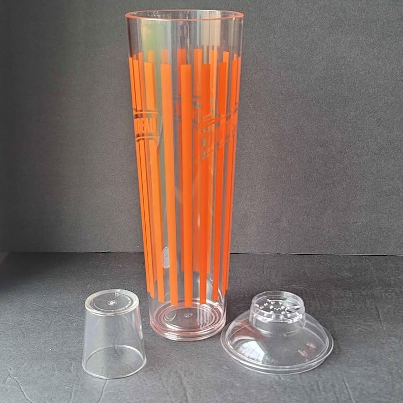 May include: A clear plastic cocktail shaker with orange vertical stripes and text. The shaker is accompanied by a small clear cup and a clear lid with a strainer. The set is designed for mixing drinks.