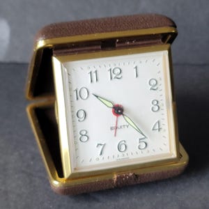 May include: A brown and gold travel alarm clock with a white face. The clock face has black numerals and hands, with the word "EQUITY" printed below the center. The clock is in a brown case that opens and closes.