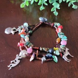 May include: A multi-strand bracelet featuring a variety of colorful beads and charms. The bracelet includes turquoise, pink, and brown beads, along with silver-toned charms of a hedgehog, a deer, and a flute player. The bracelet is displayed on a dark wooden surface.