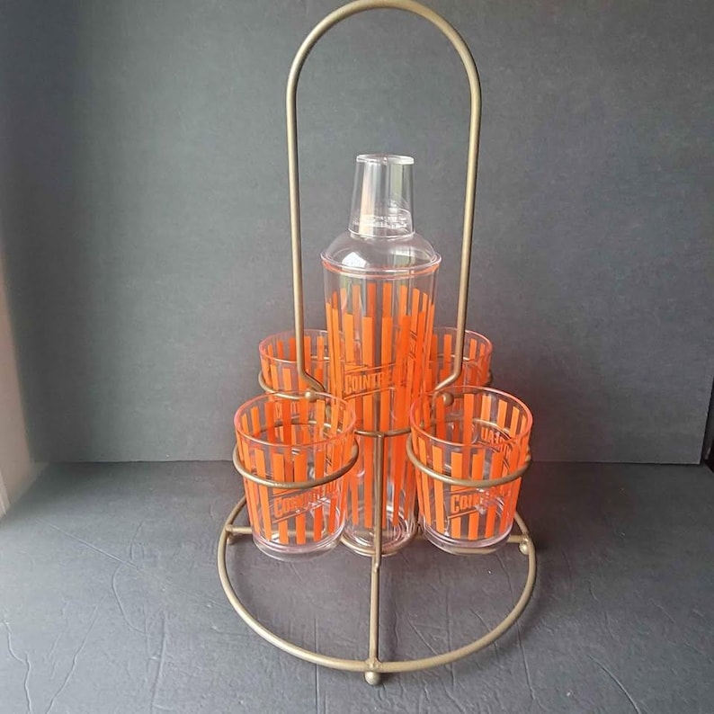 May include: Vintage cocktail shaker set with a clear shaker and six matching glasses. The glasses and shaker feature orange vertical stripes and the word "Cointreau". The set is held in a gold-colored metal stand.