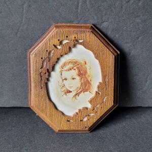 May include: A vintage portrait of a young girl in a wooden frame. The frame is octagonal with a wood grain texture and decorative cutouts. The portrait features a girl with long, light brown hair and a soft, painted style.