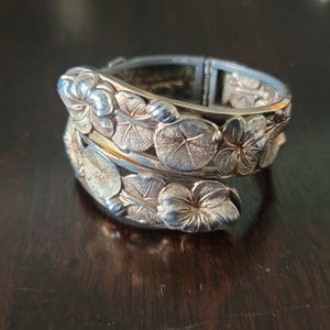 May include: Silver-toned hinged bangle bracelet with floral and leaf design. The bracelet features detailed relief carvings of flowers and leaves, creating a textured surface. The clasp is visible on the top of the bracelet.