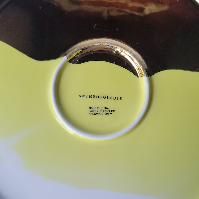 Puede incluir: Un plato de cer&aacute;mica amarillo y negro con un borde dorado. La parte inferior del plato tiene el texto "Anthropologie" y "Made in China, Fabriqu&eacute; en Chine, Handwash Only".