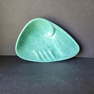 Könnte beinhalten: Ein türkisfarbener Aschenbecher im Mid-Century-Modern-Stil mit einer einzigartigen, asymmetrischen Form. Der Aschenbecher hat ein geschwungenes Design auf einer Seite und drei Rillen zum Ablegen von Zigaretten. Die gesprenkelte Glasur verleiht dem Keramikstück Textur.