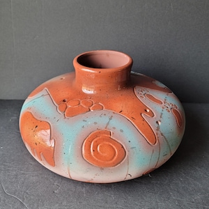 May include: A terracotta vase with a wide, rounded base and a narrow neck. The vase is decorated with a swirling pattern of blue and white glaze, and has a textured surface.