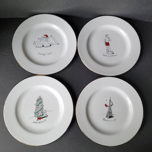 Puede incluir: Juego de cuatro platos de cerámica blanca con borde dorado. Cada plato presenta una ilustración en blanco y negro y texto. Las ilustraciones incluyen un árbol de Navidad, la Torre Eiffel, una esfinge y una estatua. El texto en los platos dice "Merry Be the Gift", "Ooh La La", "Sandy Claws", y "Back in the Ottoman".