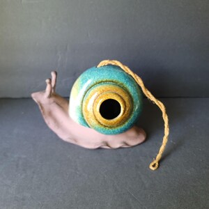 May include: A ceramic snail figurine with a smooth, matte light brown body and two antennae. The shell is a glossy, rounded form featuring a gradient of teal, blue, and golden-brown glaze with a central circular opening and a tan jute rope attached.