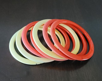 Può includere: Un mucchio di braccialetti di plastica colorati in tonalità crema, giallo e rosso. I braccialetti sono disposti in cerchio, con i braccialetti rossi all'esterno e i braccialetti crema all'interno.
