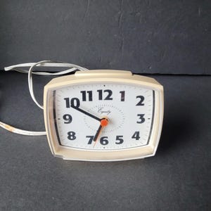 May include: A vintage cream-colored Equity brand electric alarm clock. The clock has a rectangular face with a white dial, black numerals, and a red second hand. The clock is plugged in with a white cord.