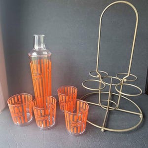 May include: A vintage cocktail set featuring a clear shaker with orange vertical stripes and the word "Cointreau". Includes four matching glasses and a gold-colored metal stand. The glasses and shaker have orange stripes.