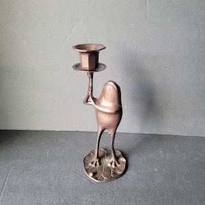 May include: A bronze-colored frog-shaped candle holder. The frog stands on a lily pad base, holding a hexagonal candle cup. The candle holder is made of metal and has a vintage appearance.