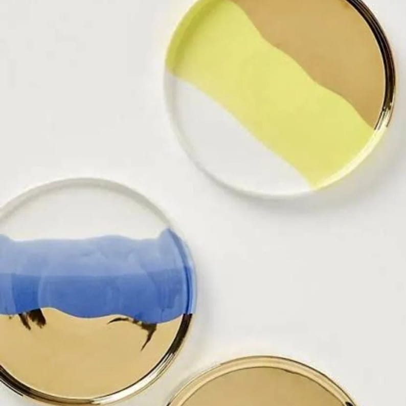 Puede incluir: Tres posavasos redondos de cer&aacute;mica con bordes dorados. Los posavasos est&aacute;n pintados con un solo color en la mitad superior y oro en la mitad inferior. Los colores son amarillo, azul y blanco.