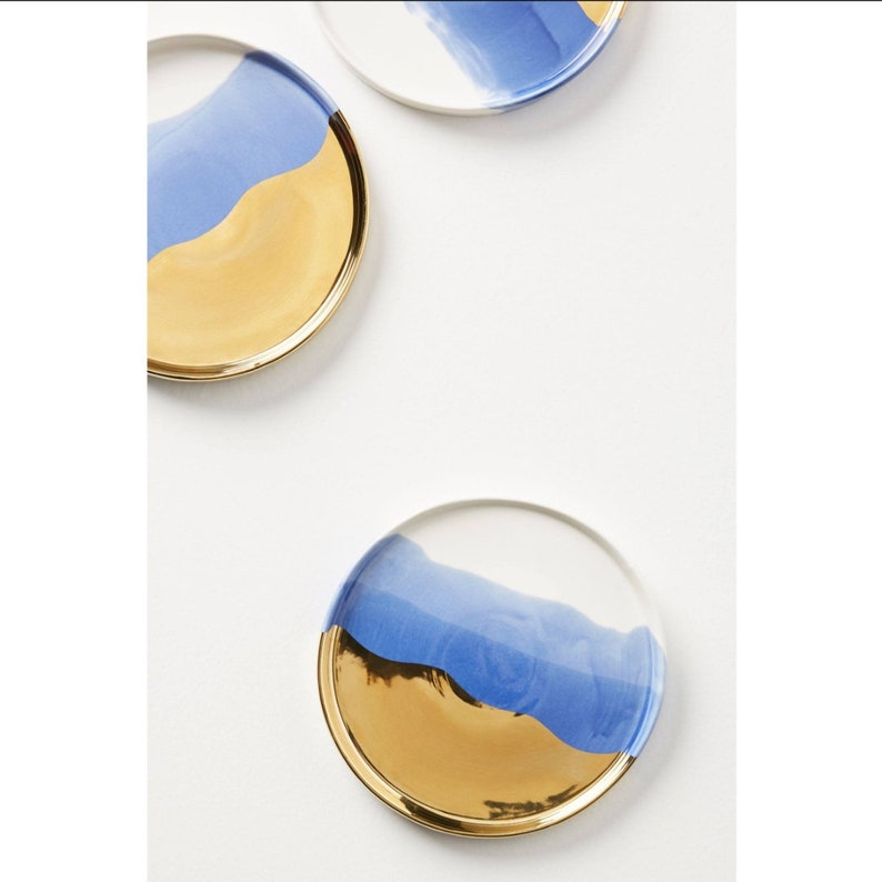 Puede incluir: Tres posavasos redondos de cer&aacute;mica con un dise&ntilde;o azul y dorado. Los posavasos tienen una base blanca con una franja azul similar a la acuarela y un borde dorado.