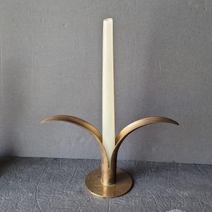 May include: A brass candle holder with a single candle in the center. The holder has three arms that curve upwards and outwards, resembling a stylized flower.
