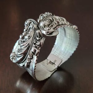 May include: Silver-toned cuff bracelet with ornate, detailed floral and face designs. The hinged bracelet features a textured band and a secure clasp. The intricate carvings and polished finish give it a vintage or antique aesthetic.