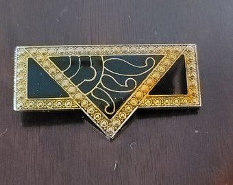 Könnte beinhalten: Rechteckige Brosche mit schwarzem Emaille-Mittelteil und goldfarbener Umrandung. Das Design zeigt ein stilisiertes Sonnenmotiv in einem dreieckigen Abschnitt. Die Ränder sind mit kleinen, kreisförmigen Akzenten versehen.