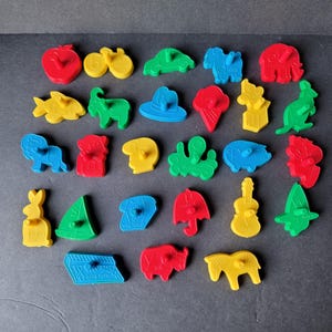 Puede incluir: Una colección de cortadores de galletas de plástico de colores en varias formas, incluyendo una manzana, un coche, un elefante y una guitarra. Los cortadores son de color rojo, amarillo, azul y verde, y están dispuestos sobre una superficie oscura.