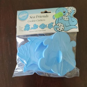 Puede incluir: Un juego de cuatro cortadores de galletas de plástico azul con forma de criaturas marinas, incluyendo un pulpo, un delfín y un pez. El embalaje dice "Sea Friends Cookie Cutters" e incluye una ilustración de dibujos animados de una criatura marina azul sosteniendo galletas.