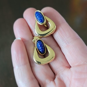 May include: A pair of gold-toned earrings with a teardrop design. Each earring features a dark blue oval stone with carved details, set within a gold frame. The earrings are displayed against a blurred background.