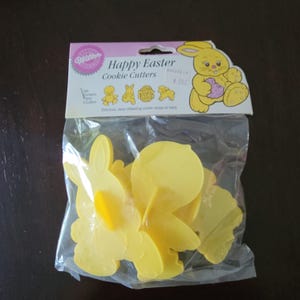 Puede incluir: Un paquete de cortadores de galletas de Pascua de plástico amarillo. El paquete incluye un conejito, un pollito, un huevo y un cordero. El embalaje dice "Happy Easter Cookie Cutters" y presenta un conejito de dibujos animados sosteniendo un huevo.