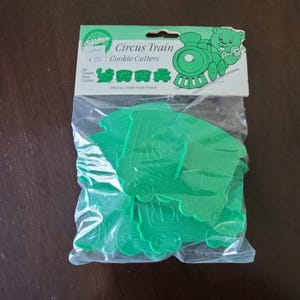 Puede incluir: Un paquete de cortadores de galletas de plástico verde con forma de tren de circo. El paquete incluye un encabezado con las palabras "Circus Train Cookie Cutters" y un gráfico de un tren y un oso. El juego contiene cuatro cortadores.
