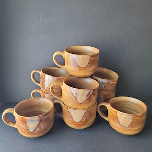 Peut inclure: Plusieurs tasses en céramique faites à la main. Les tasses sont de couleur marron chaud avec un accent plus clair, crème, sur le devant. Chaque tasse a une anse arrondie et une large ouverture, idéale pour le café ou le thé.