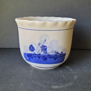 May include: A white ceramic planter with a blue Dutch windmill scene. The planter has a scalloped rim and a blue band around the top. The windmill scene depicts a windmill, trees, and houses. The planter is approximately 5 inches tall.