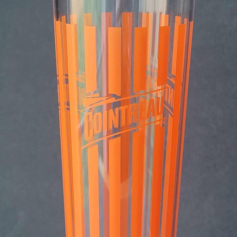 May include: A tall, clear glass with vertical orange stripes. The word "COINTREAU" is printed in orange on the glass. The glass is designed for cocktails and other beverages.