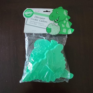 Puede incluir: Un paquete de cortadores de galletas de dinosaurios de plástico verde. El paquete incluye una ilustración de dinosaurio de dibujos animados y el texto "Dinosaur Cookie Cutters". Los cortadores están en una bolsa de plástico transparente.