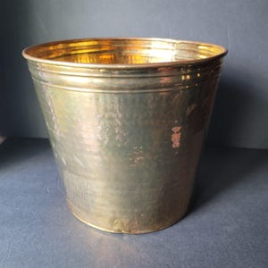 May include: A hammered brass bucket with a slightly tapered shape. The bucket has a wide, rounded rim and a textured surface. The brass has a warm, golden hue, reflecting light. The bucket is empty and sits on a dark surface.