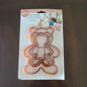 Puede incluir: Un paquete de cortadores de galletas Wilton Bear Family. El paquete contiene cuatro cortadores de galletas con forma de oso de diferentes tamaños. Los cortadores son de color naranja claro y están hechos de plástico. El embalaje tiene un fondo blanco con ilustraciones de galletas de oso decoradas.