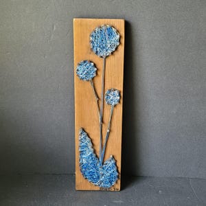 May include: Wooden wall art featuring a string art floral design. The artwork has three blue and white flower heads and a blue leaf, all made with string. The flowers are on thin metal stems, and the piece is mounted on a rectangular wooden board.