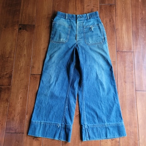 May include: Vintage blue denim pants with wide legs and large front pockets. The pants have a button fly and show signs of wear, including a small tear on the front pocket. The denim is a medium wash, and the pants have a high waist.