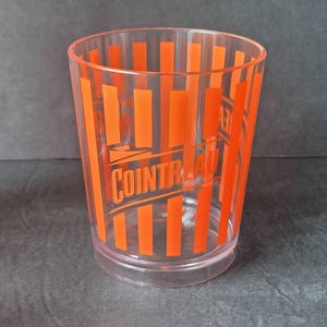 May include: A clear plastic tumbler with vertical orange stripes and the word "COINTREAU" printed in orange. The glass is cylindrical with a slightly flared top and a clear base.
