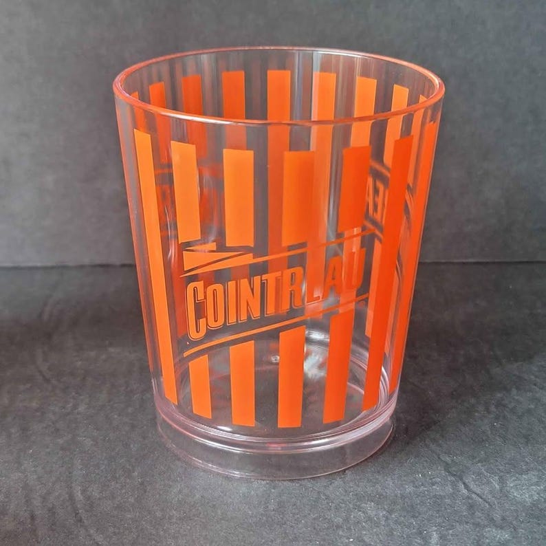May include: A clear plastic tumbler with vertical orange stripes and the word "COINTREAU" printed in orange. The glass is cylindrical with a slightly flared top and a clear base.