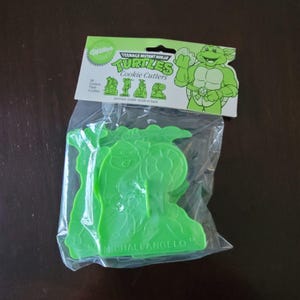 Puede incluir: Un paquete de cortadores de galletas de plástico verde de las Tortugas Ninja. El paquete incluye cuatro cortadores, con el nombre "MICHAEL ANGELO" impreso en uno de los cortadores. El embalaje también presenta el logotipo "Wilton" y las palabras "Cookie Cutters".