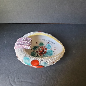 May include: Hand-painted ceramic ashtray with a floral design. The ashtray is white with red, blue, and green accents. The rim is decorated with small white dots and a yellow border. The text "SELVA DI FASANO" is written on the inside.