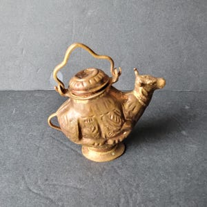 May include: A small, antique brass camel-shaped teapot with a handle and a lid. The teapot has intricate embossed details and sits on a round base. The camel's head is raised, and the spout is at the front.