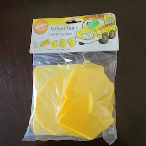 Puede incluir: Un paquete de cortadores de galletas de plástico amarillo en varias formas, incluyendo un autobús escolar, una manzana y un boleto. El paquete está etiquetado como "School Days Cookie Cutters" con una ilustración de autobús escolar.
