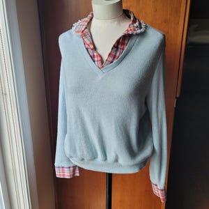 Op de afbeelding: Een lichtblauwe V-hals trui met lange mouwen, gelaagd over een geruit overhemd met kraag. De kraag en manchetten van het overhemd hebben een rood, wit en blauw ruitpatroon. De trui wordt op een mannequin getoond.