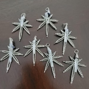 Op de afbeelding: Zes zilverkleurige metalen hangers in de vorm van zevenpuntige bladeren, elk voorzien van een heldere, verticale kristalpunt die aan de bovenkant met draad is omwikkeld. Elke hanger heeft een klein rond oogje voor bevestiging aan een ketting.