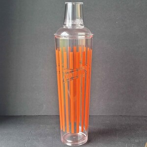 May include: A clear cocktail shaker with a transparent lid and a base. The shaker features vertical orange stripes and the word "COINTREAU" printed in the center. The shaker is designed for mixing drinks.