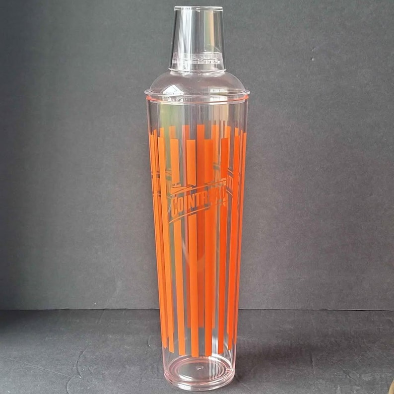 May include: A clear cocktail shaker with a transparent lid and a base. The shaker features vertical orange stripes and the word "COINTREAU" printed in the center. The shaker is designed for mixing drinks.