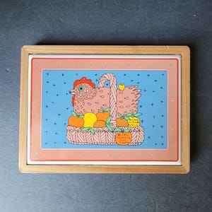 May include: A rectangular ceramic trivet with a wooden frame. The trivet features a pink chicken in a basket filled with oranges and apples, with a small yellow chick perched on the basket handle. The background is blue with small white dots.