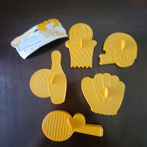 Puede incluir: Un conjunto de cortadores de galletas amarillos con temática deportiva, que incluyen una canasta de baloncesto, un casco de fútbol americano, un guante de béisbol, un bolos y una raqueta de tenis. También se incluye un paquete con texto.