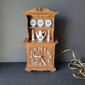 May include: A vintage wooden clock with a hutch-style top. The top shelf displays three small plates with blue floral designs, a teapot, and two candle holders. The clock face has white numerals and hands. A power cord extends from the back.