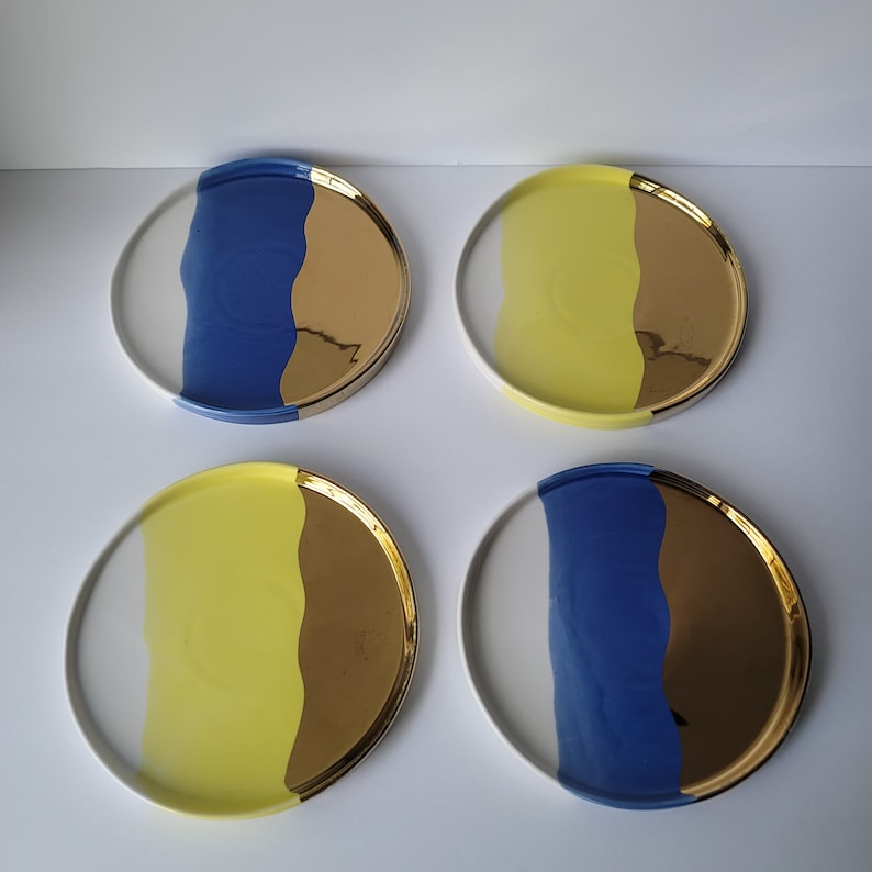 Puede incluir: Juego de cuatro platos de cer&aacute;mica con un dise&ntilde;o blanco, amarillo, azul y dorado. Cada plato tiene una l&iacute;nea ondulada que divide los colores.