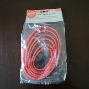 Puede incluir: Un paquete de cortadores de galletas ovalados Wilton. Los cortadores son de plástico naranja y de varios tamaños. El embalaje incluye el texto "Oval Cookie Cutters" y "Versatile! Use as cookie cutters and as fast and easy pattern press-in cubes."