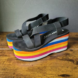 May include: Black platform sandals with colorful rainbow striped wedge heels. The sandals have black straps and the brand name "KET DOG" is visible on the footbed. The platform heels have a white sole and are approximately 3 inches high.