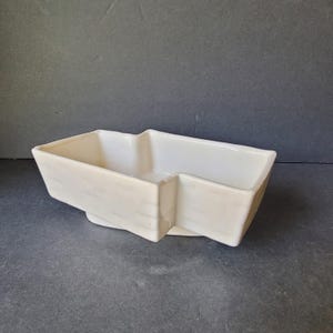 Könnte beinhalten: Ein cremefarbener, rechteckiger Keramik-Pflanztopf mit einer einzigartigen sternartigen Form. Der Pflanztopf hat einen erhöhten Sockel und abgewinkelte Seiten, wodurch ein modernes und geometrisches Design entsteht. Ideal für Sukkulenten oder kleine Pflanzen.